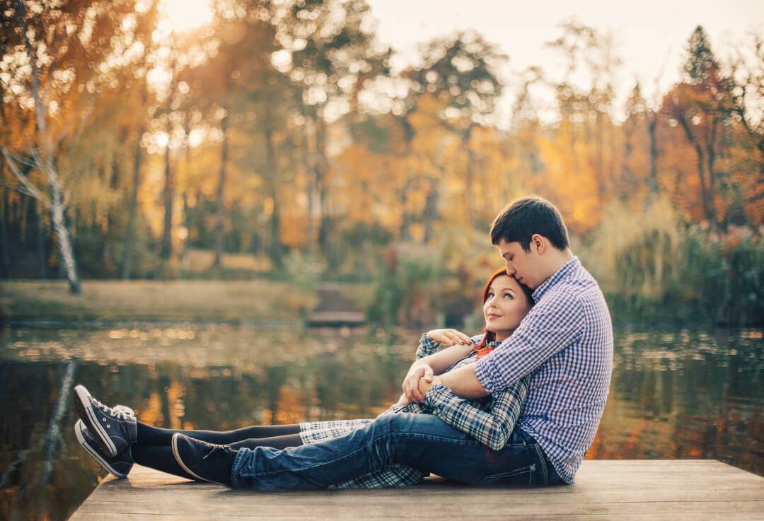 Husband holding his wife affectionately while sitting on a dock.